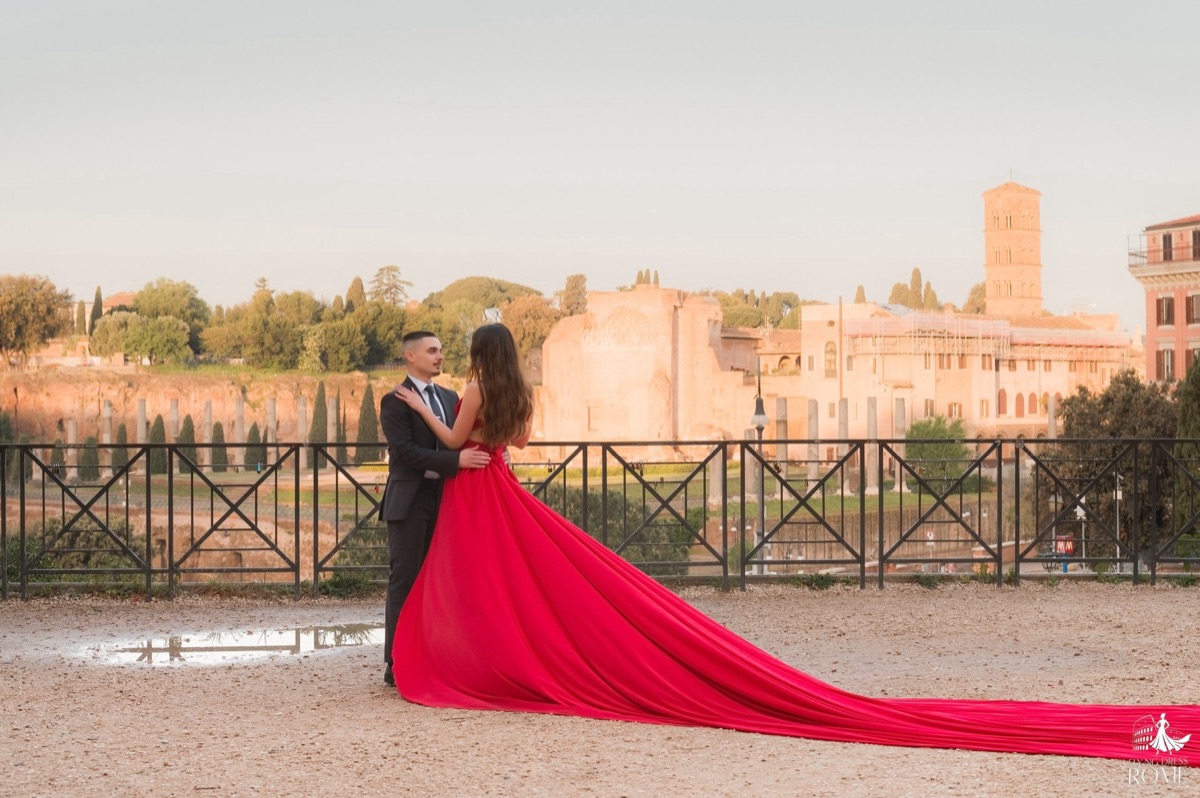 Couple flying dress pose The View: couple standing still almost embracing, her back to the camera, red train stretched out on the ground, Roman Forum, Rome