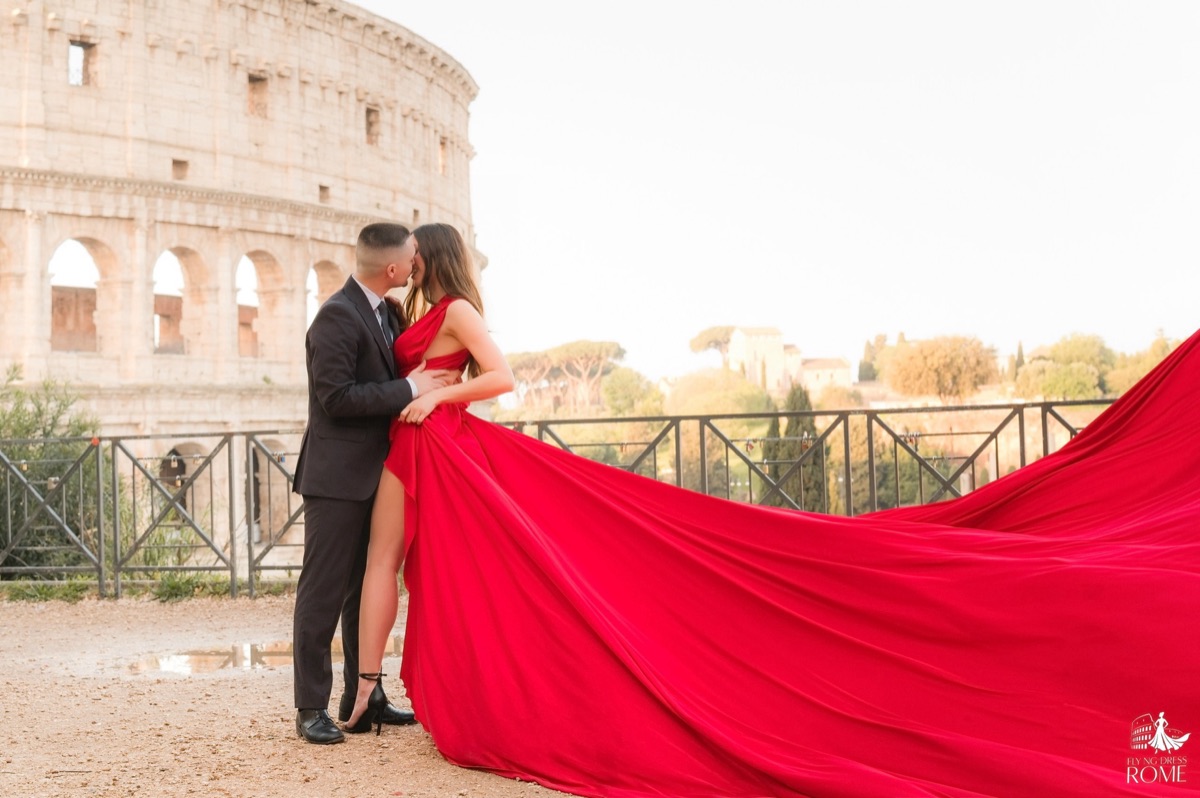 Couple flying dress pose The Embrace: he holds her at the waist, red train thrown behind them, Colosseum at sunrise, Rome